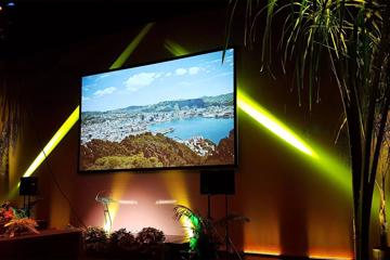 A stage setup at St James Theatre, in Te Aro, Wellington, with decorative foliage, bright spotlights, a couple of speakers and an atrial image of Wellington projected onto a large screen.