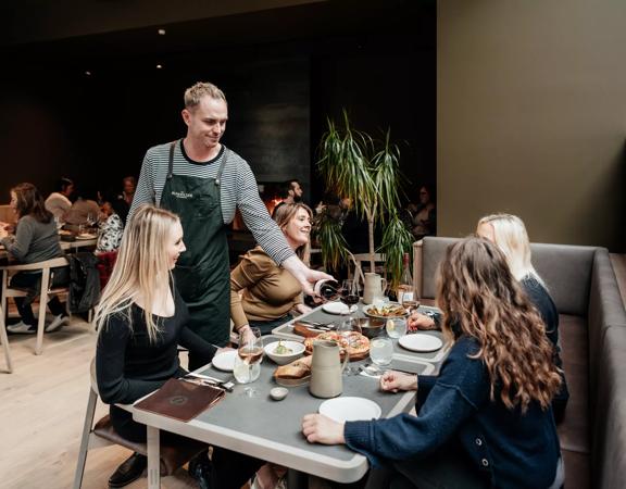 bartender pouring a glass of red wine to 4 customers sitting down and enjoying their food and drinks inside the Runholder.