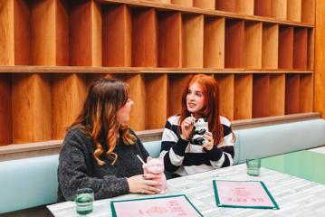 Two friends chat over drinks at Mr. Go's in Wellington. They're sitting next to each other on a pale blue bench along a decorative wooden wall and their drinks are served in animal-shaped cups.