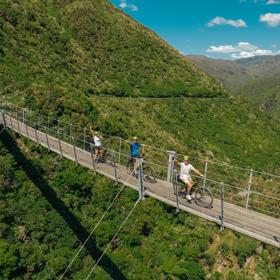 Aerial shot of mountain bikers on a swing bridge above a green valley on the Remutaka Cycle Trail.
