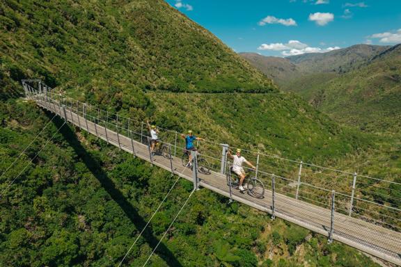 Aerial shot of mountain bikers on a swing bridge above a green valley on the Remutaka Cycle Trail.

