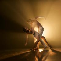 Two Royal New Zealand Ballet dancers illuminated by warm amber light, performing in a spacious, minimalist setting.