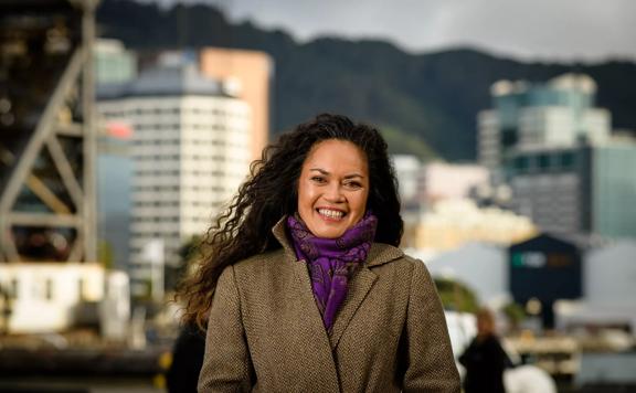 Director Ngā Toi Māori for the Aotearoa New Zealand Festival of the Arts, Mere Boynton, wearing a brown coat and purple scarf, standing and smiling at the Wellington waterfront.