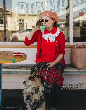 A chic coffee drinker sits outside of Queen Sally's Diamond Deli with two small dogs.