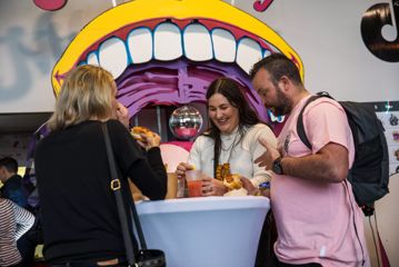 3 people standing at a bar table eating hotdogs and drinking beer with colourful pop art of a mouth in the background.
