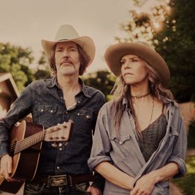 Gillian Welch & David Rawlings in cowboy hats standing outdoors, one holding an acoustic guitar.