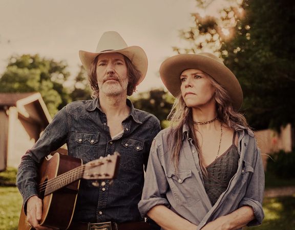 Gillian Welch & David Rawlings in cowboy hats standing outdoors, one holding an acoustic guitar.