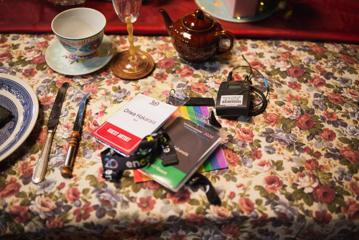 A table with a floral tablecloth, a teapot, a teacup, a wine glass and printed pass lanyards.