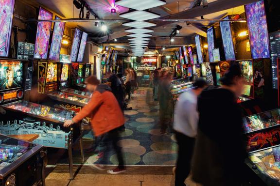 Looking straight down the middle of the arcade with pinball machines on either side. Multiple neon lights and blinking lights from the machines. There are several people playing arcade games, some of whom are blurred in mid movement.