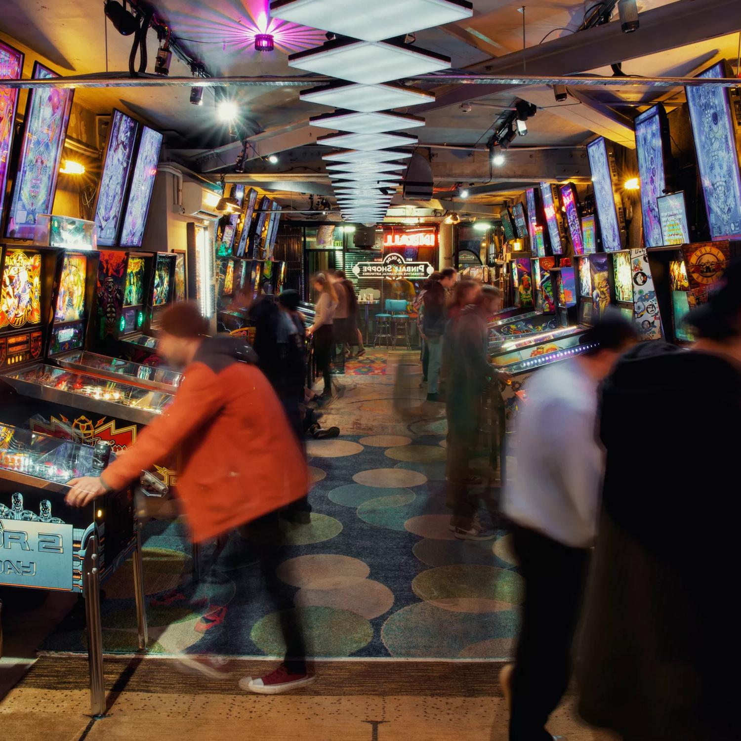 Looking straight down the middle of the arcade with pinball machines on either side. Multiple neon lights and blinking lights from the machines. There are several people playing arcade games, some of whom are blurred in mid movement.