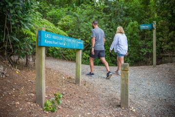 The people walk along a wide gravel path passing the entrance sign to East Harbour Regional Park.