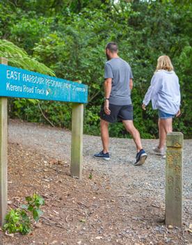 The people walk along a wide gravel path passing the entrance sign to East Harbour Regional Park.