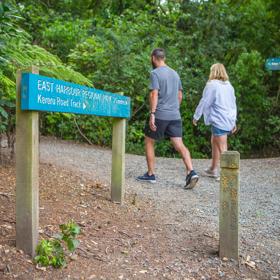 The people walk along a wide gravel path passing the entrance sign to East Harbour Regional Park.