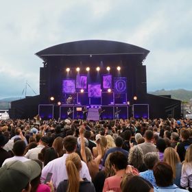 A large crowd watches a live music performance on an outdoor stage, with giant screens on either side and hills visible in the background.
