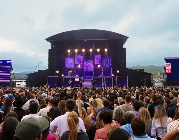 A large crowd watches a live music performance on an outdoor stage, with giant screens on either side and hills visible in the background.