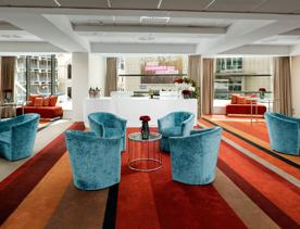 The hotel bar at The Rydges Hotel in Wellington. The bar is in the  centre background of the image, set up with champagne flutes, wine glasses and bottles of wine. The carpet is striped and there are five small tables with a variety of seating options.