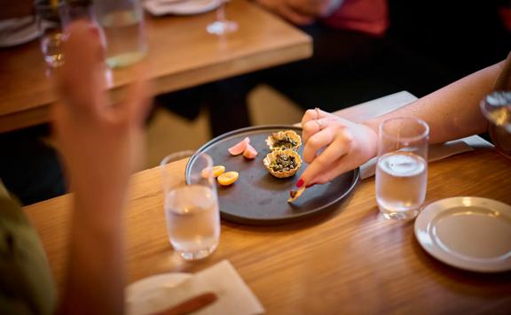 An entré dish comprised of small shared snacks sits between two water glasses on a table. A hand with red-painted nails sneaks in for a sample.