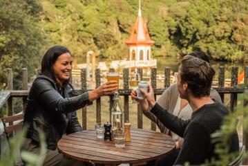 Three people sit at a small wooden table on a deck clicking glasses. They overlook the water tower at Zealandia.