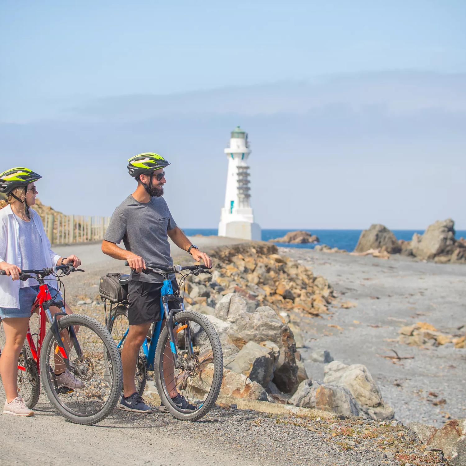 2 people standing still with their bikes and helmets on, staring out into the ocean. The Pencarrow light house and large rocks can be seen behind them