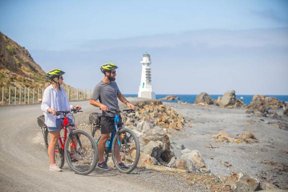2 people standing still with their bikes and helmets on, staring out into the ocean. The Pencarrow light house and large rocks can be seen behind them