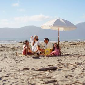 A family sits on Raumati beach on the sand under an umbrella. Kapiti Island is in the background.