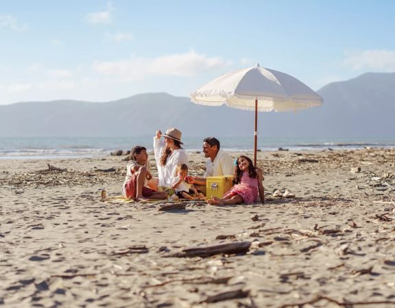 A family sits on Raumati beach on the sand under an umbrella. Kapiti Island is in the background.