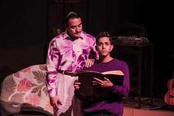 Two actors on stage, one has their hand on a younger actors shoulder as they read a large book.