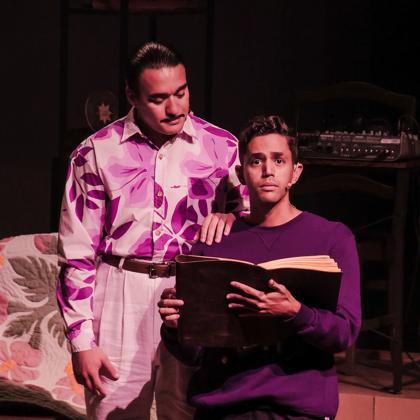 Two actors on stage, one has their hand on a younger actors shoulder as they read a large book.
