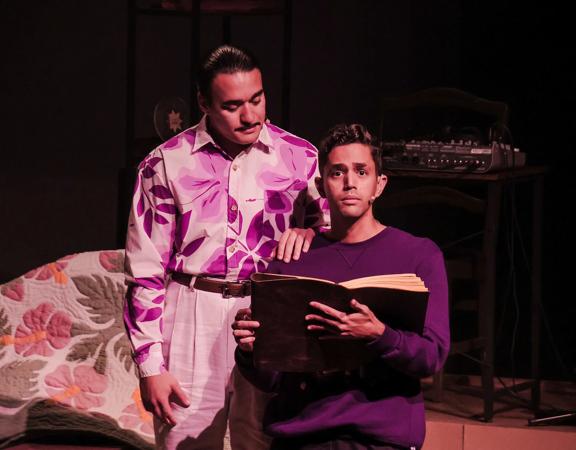 Two actors on stage, one has their hand on a younger actors shoulder as they read a large book.