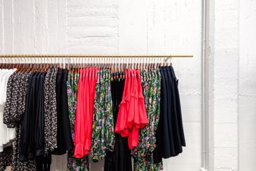 A clothing rack with floral, navy blue, coral, black and white blouses and dresses in front of a white brick wall.