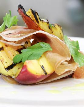 Super close up of a plate of food, containing a slightly charred nectarine with greens and prosciutto.