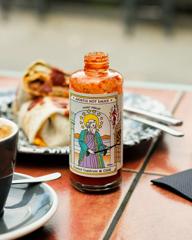 A bottle of Apostle’s roasted capsicum & chilli hot sauce with a plate of food and a coffee on a red-tiled table top.