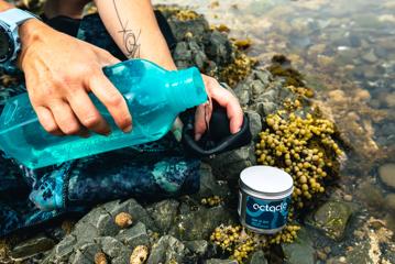 Water is being poured from a bright blue bottle onto a wetsuit on a rocky beach with a tin of Octacle wetsuit lube in the foreground.