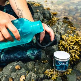 Water is being poured from a bright blue bottle onto a wetsuit on a rocky beach with a tin of Octacle wetsuit lube in the foreground.