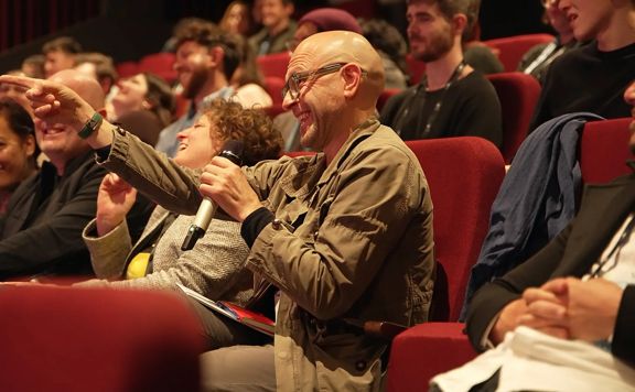 Audience member seated in a red cinema chair raises a hand while participating in a discussion at Pitchfest 2025 at Roxy Cinema.