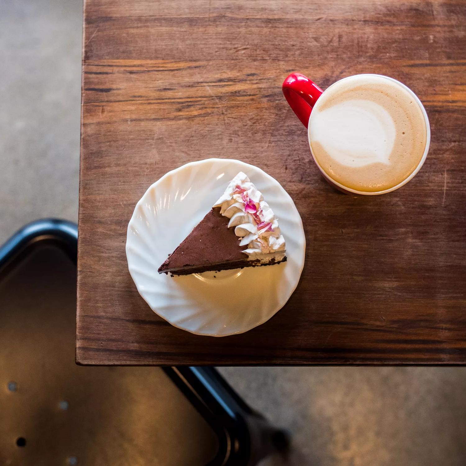 A slice of chocolate cake with white frosting and a flat white latte in a red mug on a wood table at Sweet Release, a plant-based bakery in Te Aro, Wellington.