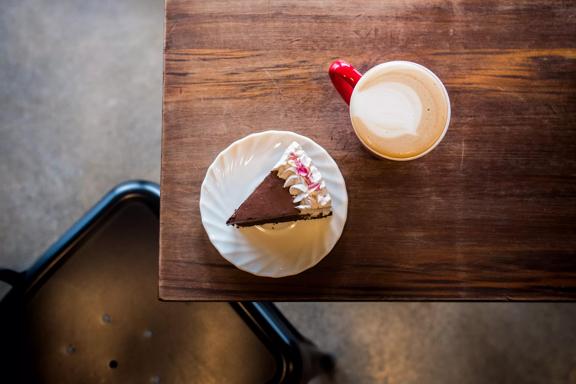 A slice of chocolate cake with white frosting and a flat white latte in a red mug on a wood table at Sweet Release, a plant-based bakery in Te Aro, Wellington.