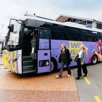 Two people walking toward a colourful minibus, covered with World of WearableArt branding.