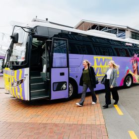 Two people walking toward a colourful minibus, covered with World of WearableArt branding.