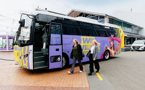 Two people walking toward a colourful minibus, covered with World of WearableArt branding.
