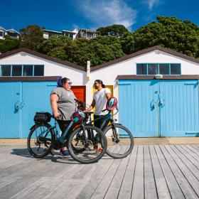 Two friends take a break from cycling in front of the colourful boatsheds at Oriental Bay in Wellington on a sunny day.