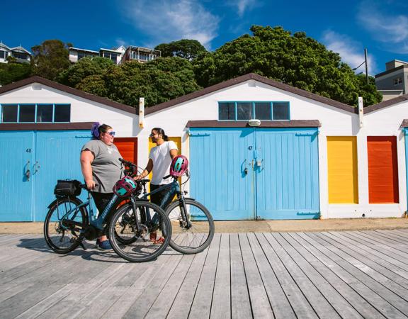 Two friends take a break from cycling in front of the colourful boatsheds at Oriental Bay in Wellington on a sunny day.