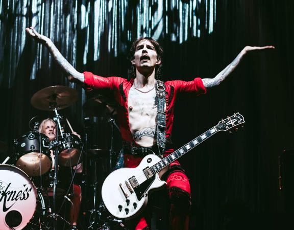 Performer from The Darkness holds hands in the air while a guitar is trapped around is waist on stage.