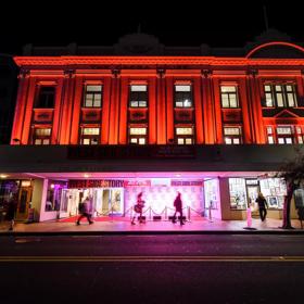 Exterior of The Opera House from the road, its night time and the building is lit up with red and pink LED lights for 'West Side Story'.