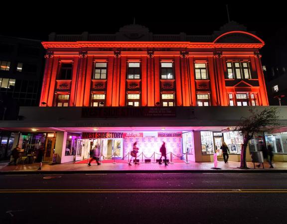 Exterior of The Opera House from the road, its night time and the building is lit up with red and pink LED lights for 'West Side Story'.