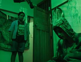 A still from the Wellington-based film, Little Apocalypse, with one person standing looking at another person who's crouching in a room with green-coloured lighting. 