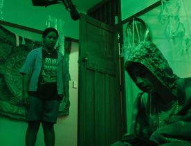 A still from the Wellington-based film, Little Apocalypse, with one person standing looking at another person who's crouching in a room with green-coloured lighting.