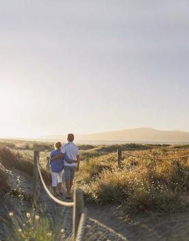 2 people walk arm in arm along the path in the sand dunes next to Waimeha Lagoon on Waikinae beach.