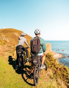 Two people on bikes on top of a grassy cliff looking out across the ocean. Pencarrow Lighthouse is in the distance.