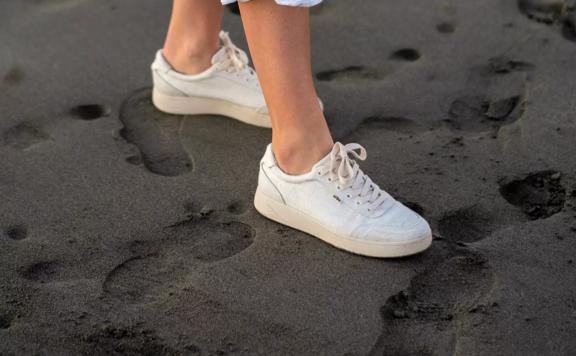 A person wearing white Orba sneakers walking in wet sand.